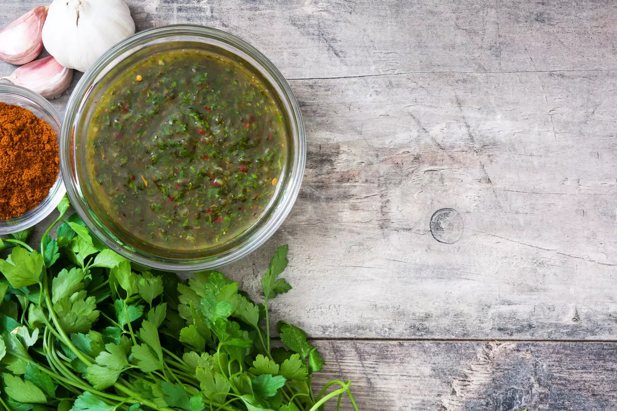 Um pote de molho chimichurri cercado por alho fresco, paprica, e um maço de salsa verde fresca, sobre uma superfície de madeira rústica para o post "o que é chimichurri".