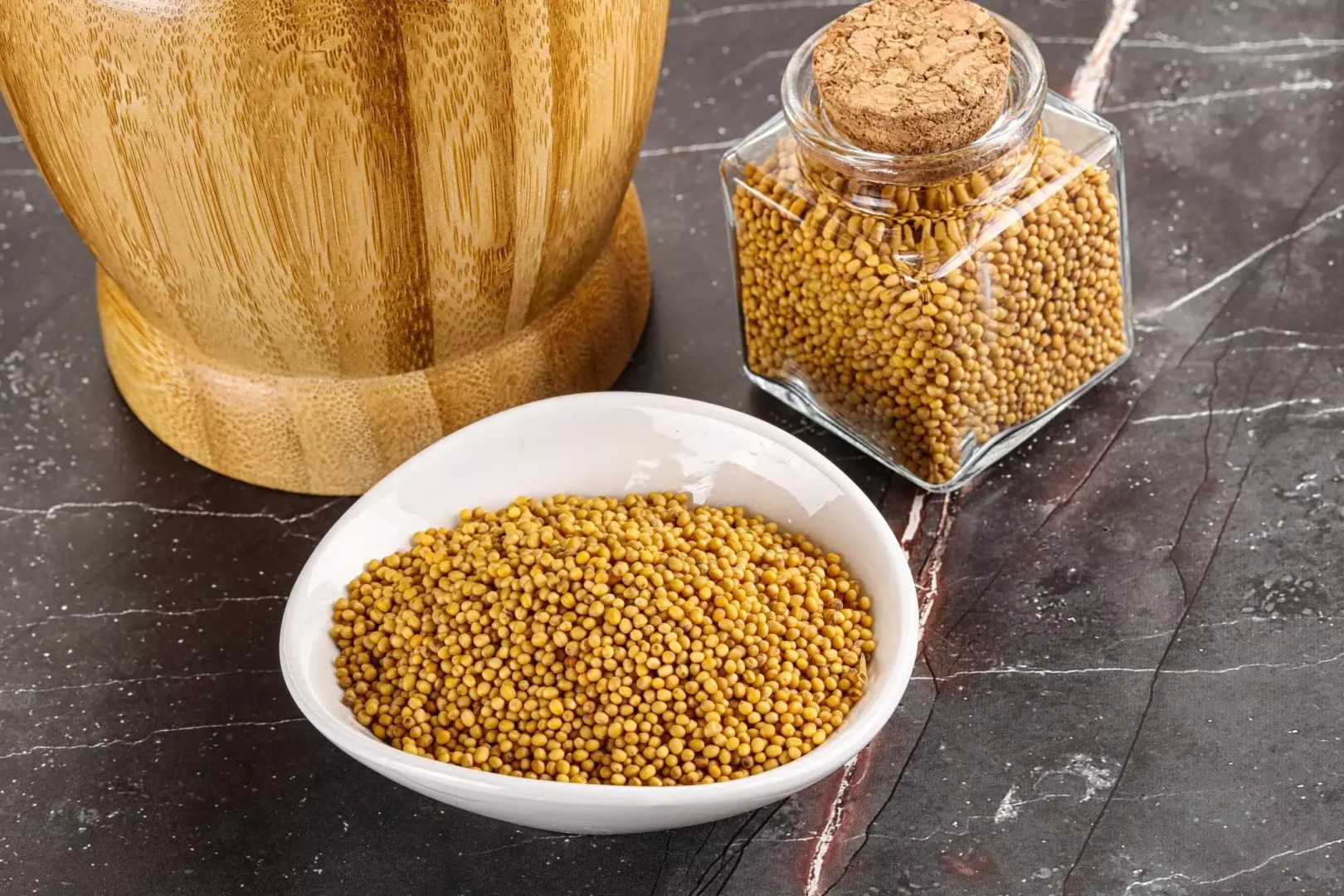 Grãos de mostarda amarela em uma tigela branca ao lado de um pote de vidro com cortiça e um moedor de madeira em fundo de mármore escuro.