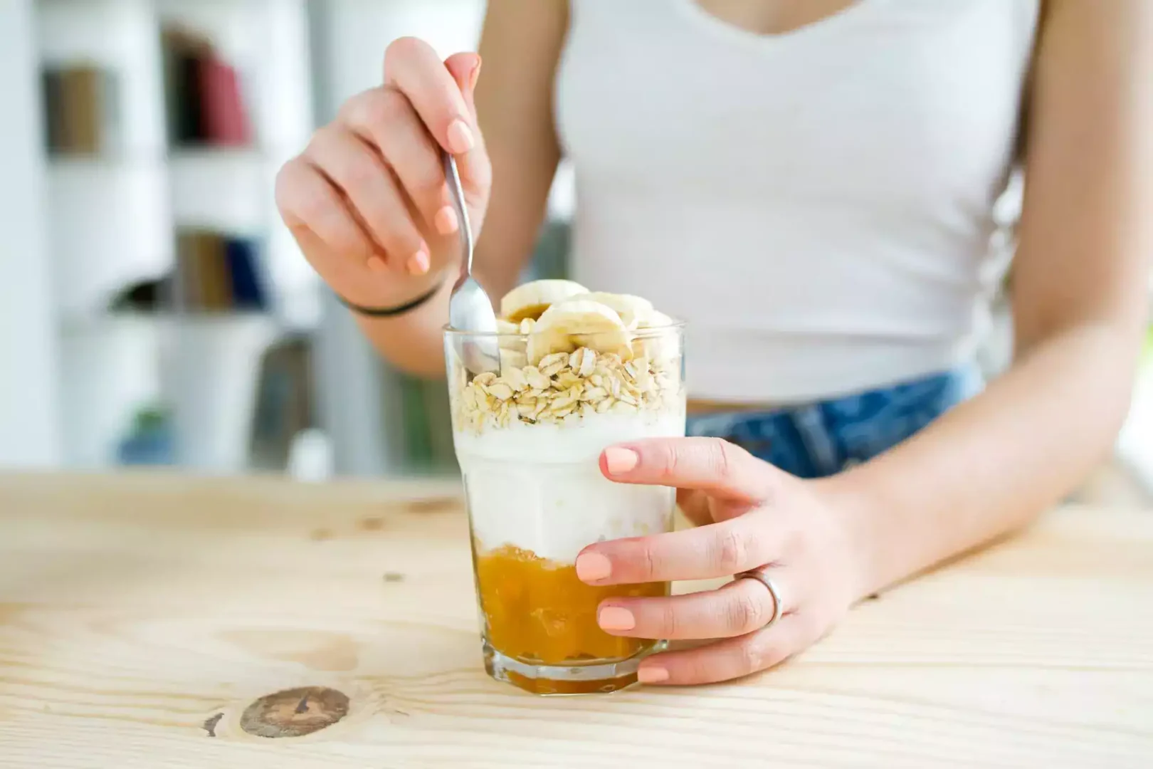 Shake de extrato de soja em um copo alto e transparente. O líquido é de cor amarelo-alaranjada vibrante, com uma textura suave e cremosa. Uma espuma densa e branca se acumula na superfície, coroando a bebida. O copo está posicionado sobre uma mesa de madeira rústica onde uma mulher segura o copo com a mão esquerda enquanto coloca uma colher com a mão direita.
