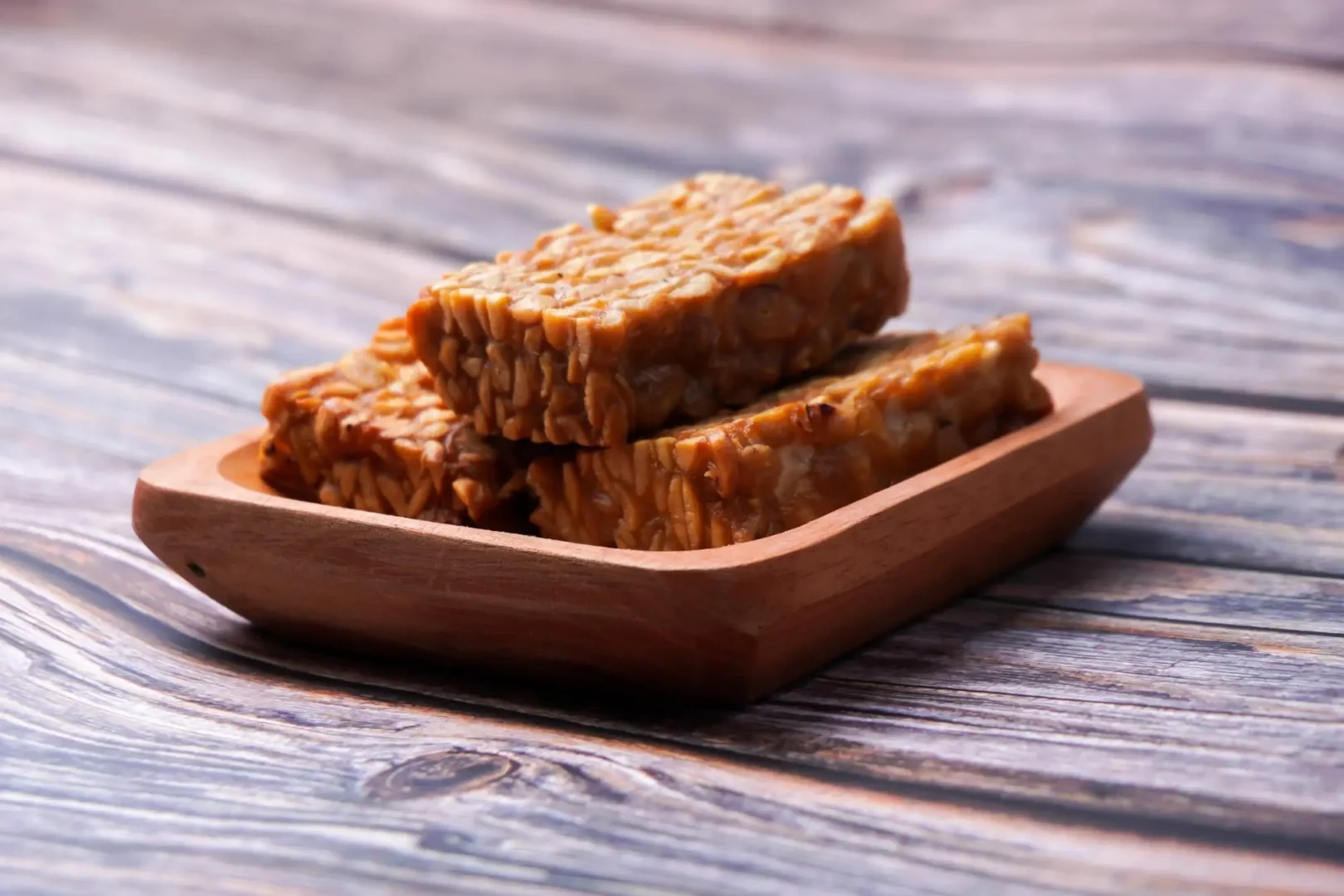 Um prato redondo de cerâmica branca, com bordas onduladas, está posicionado sobre uma mesa de madeira rústica. Dentro do prato, uma porção generosa de tempeh grelhado em tiras douradas e crocantes domina a cena.