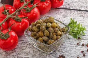 Bowl de vidro com alcaparras em primeiro plano, ao lado de galhos de tomates vermelhos com talo e folhas verdes, sobre uma superfície de madeira clara com grãos de pimenta espalhados.