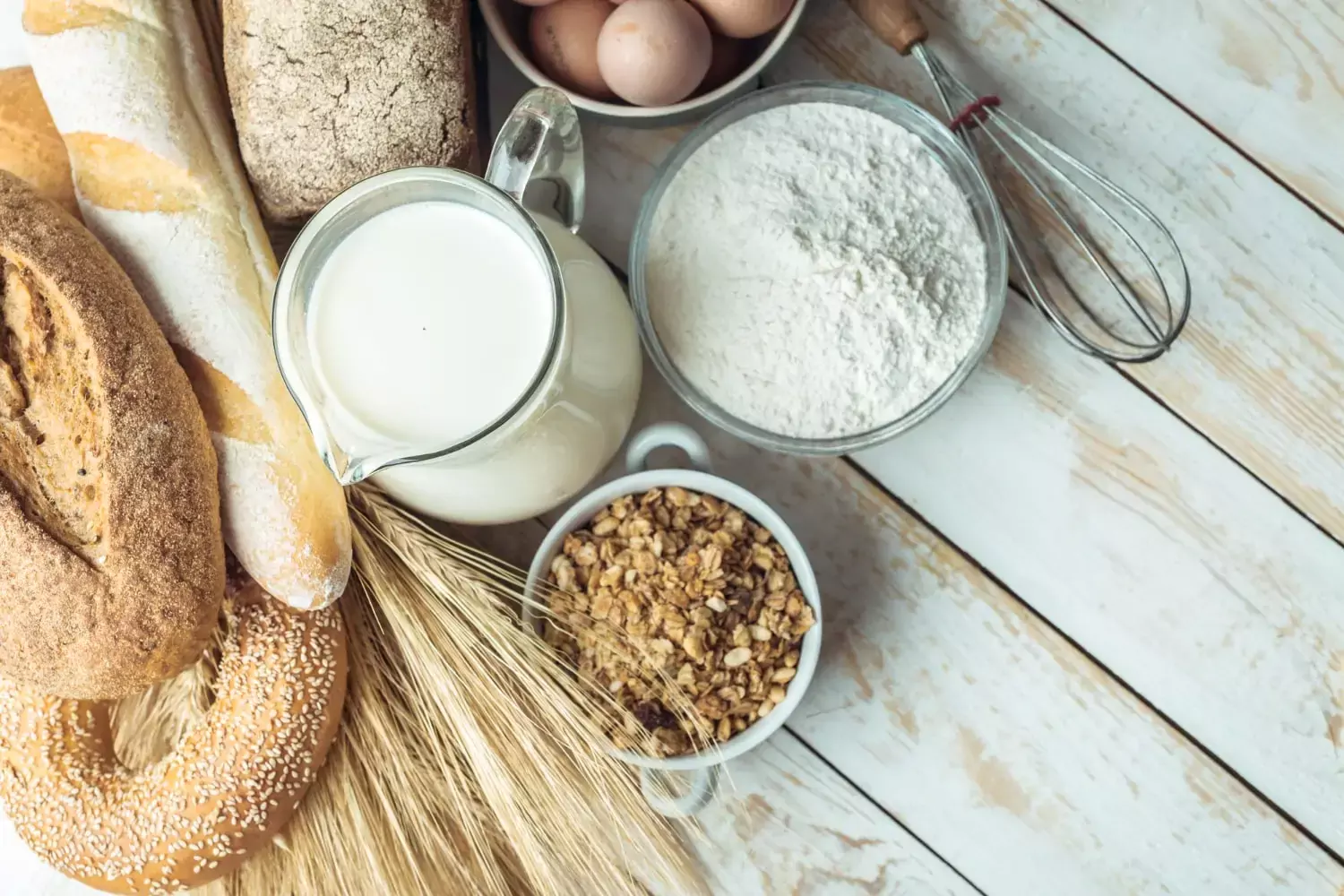 Variedade de produtos de panificação e ingredientes em uma superfície de madeira clara, incluindo pães com sementes de gergelim, jarro de leite, farinha de aveia em uma peneira, ovos, tigela de granola e espigas de trigo.