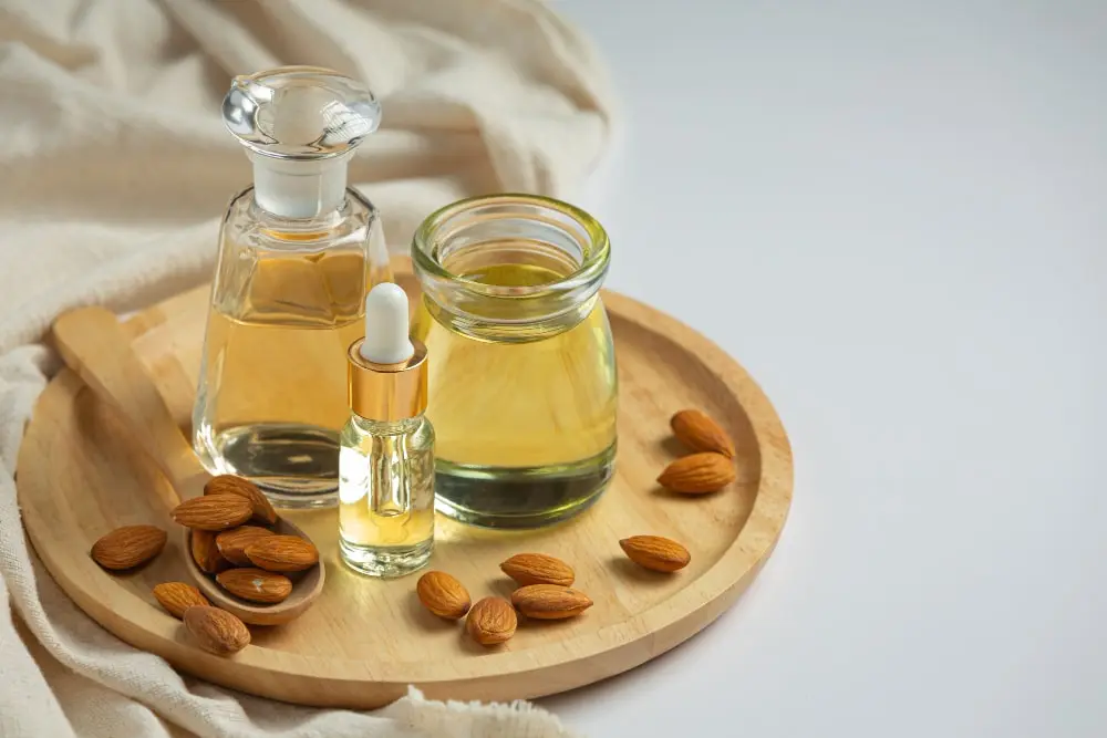 Disposição de óleo de amêndoas, com dois frascos de vidro transparente contendo o líquido dourado - um com uma tampa e outro com um conta-gotas - e um recipiente aberto, todos colocados sobre uma tábua de corte circular de madeira clara. Ao lado, há um punhado de amêndoas inteiras, que servem como indicação visual da origem do óleo. O fundo neutro com tecido bege e a iluminação natural realçam a qualidade orgânica e a pureza do produto, evocando uma sensação de bem-estar e cuidado natural.