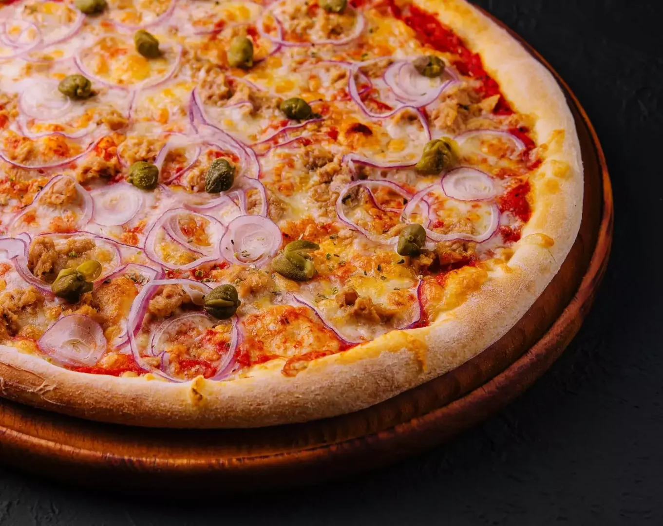 Pizza inteira com atum, cebola roxa e alcaparras em uma tábua de madeira sobre fundo preto texturizado.