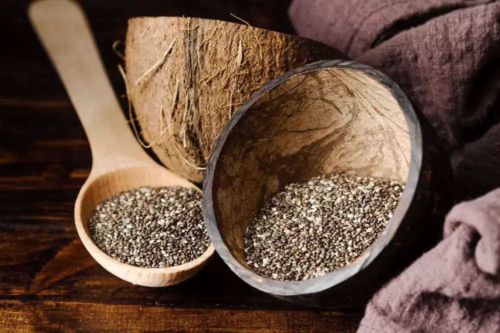 Colher de madeira e tigela de coco com sementes de chia, em um cenário rústico com iluminação suave e tecido texturizado ao fundo.