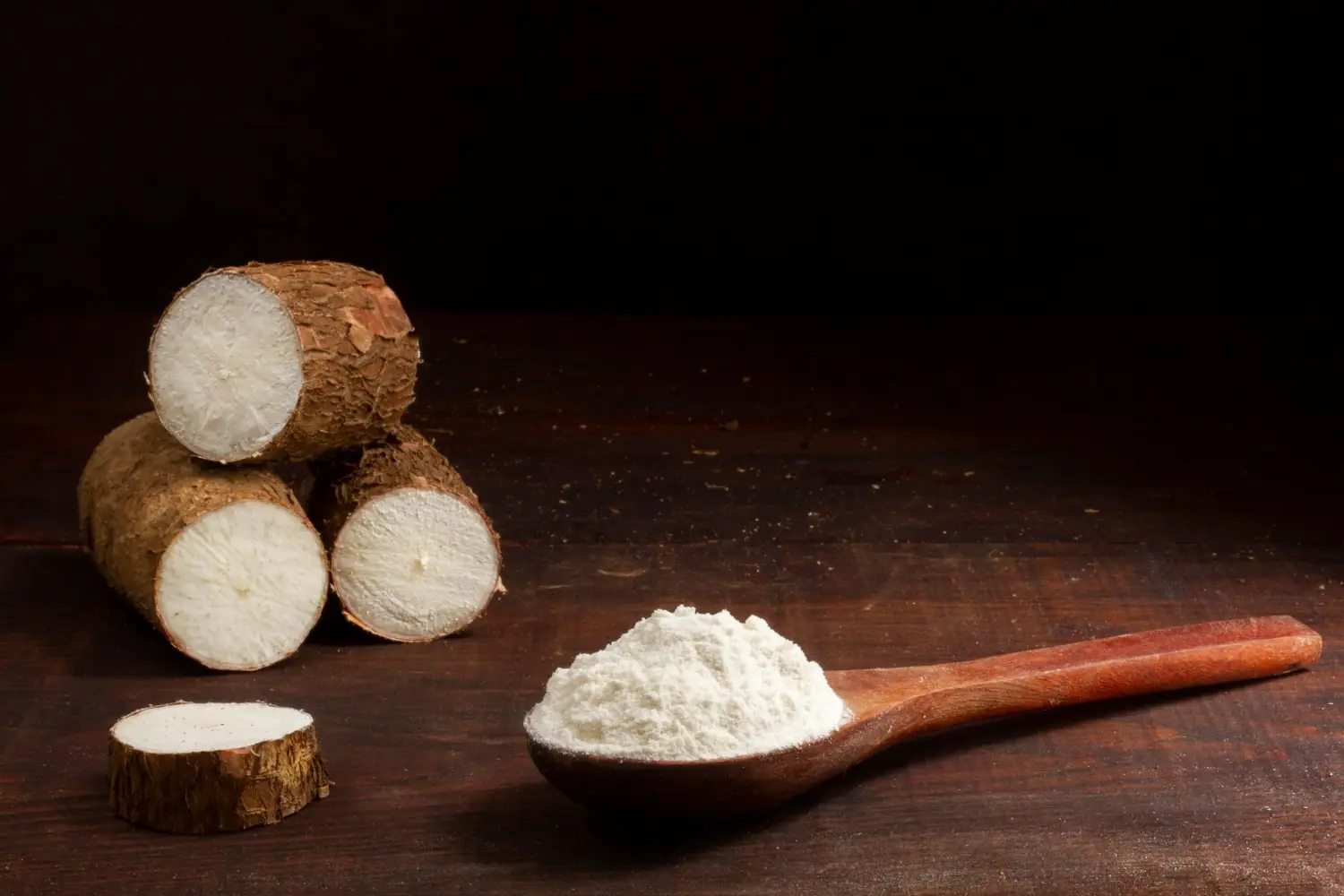 Uma composição mostrando raízes de mandioca cortadas em pedaços sobre uma superfície escura com uma colher de madeira cheia de farinha de tapioca ao lado, destacando a matéria-prima e o produto derivado