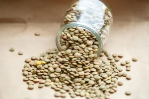 Vista de um pote de vidro tombado derramando lentilhas sobre uma superfície bege. As lentilhas, de cores variadas que vão do amarelo ao marrom, se espalham desordenadamente pela superfície, criando uma imagem rica em texturas e tons neutros.