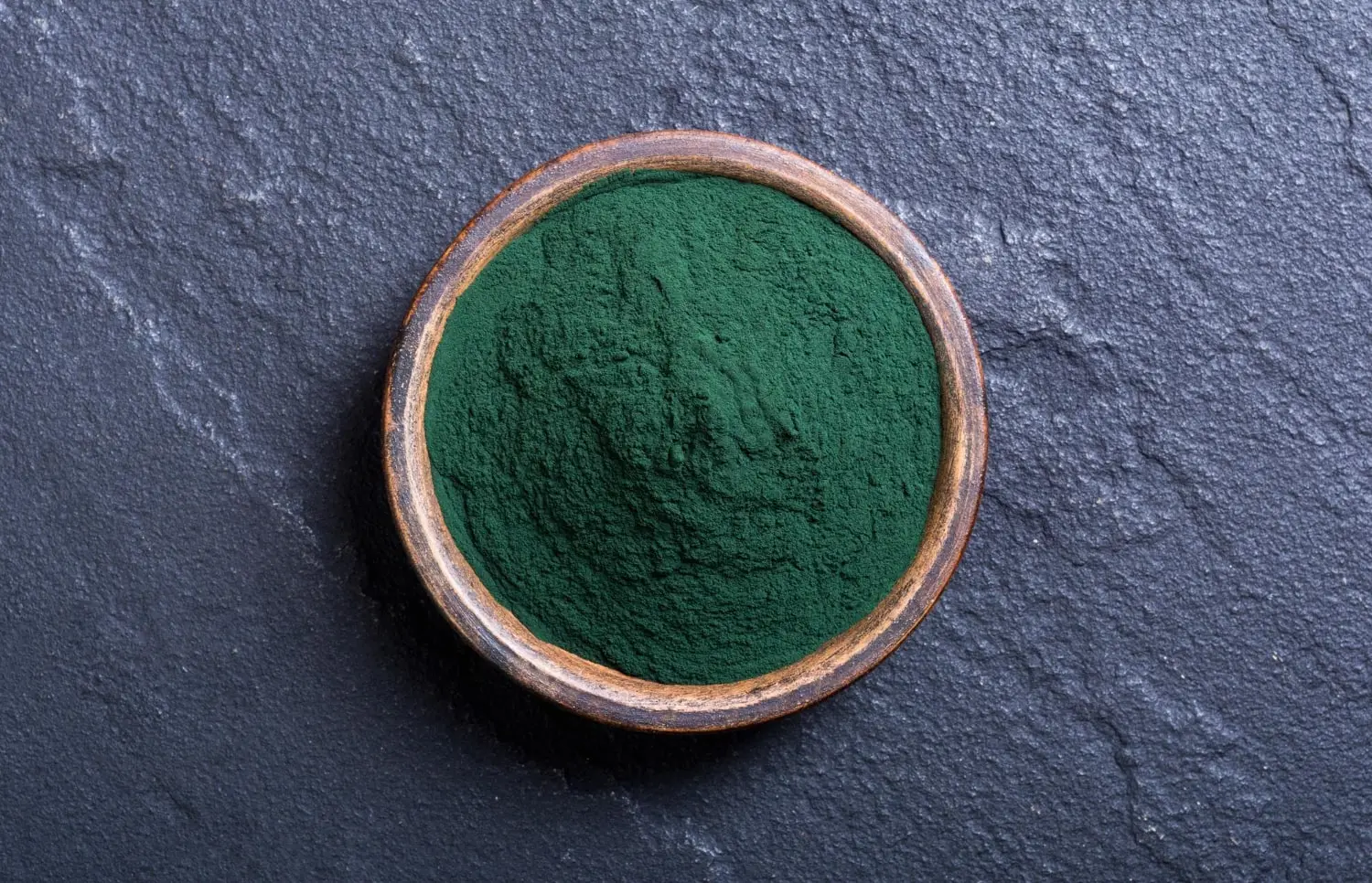 Uma tigela rústica com spirulina em pó sobre uma superfície de pedra escura.