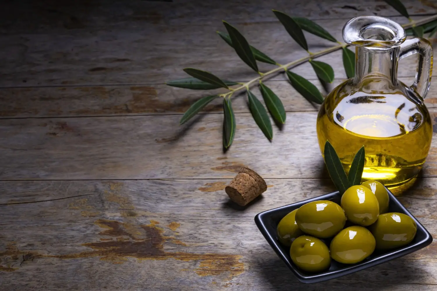 Uma jarra de azeite de oliva e uma tigela de azeitonas verdes em uma mesa de madeira. O azeite é claro e as azeitonas são brilhantes e uniformes em tamanho.