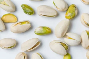 Imagem de pistaches descascados e com casca espalhados em uma superfície branca. Alguns pistaches estão parcialmente abertos, revelando a semente verde-clara dentro.