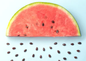 Metade de uma fatia de melancia mostrando a polpa vermelha e várias sementes espalhadas em um fundo azul claro.