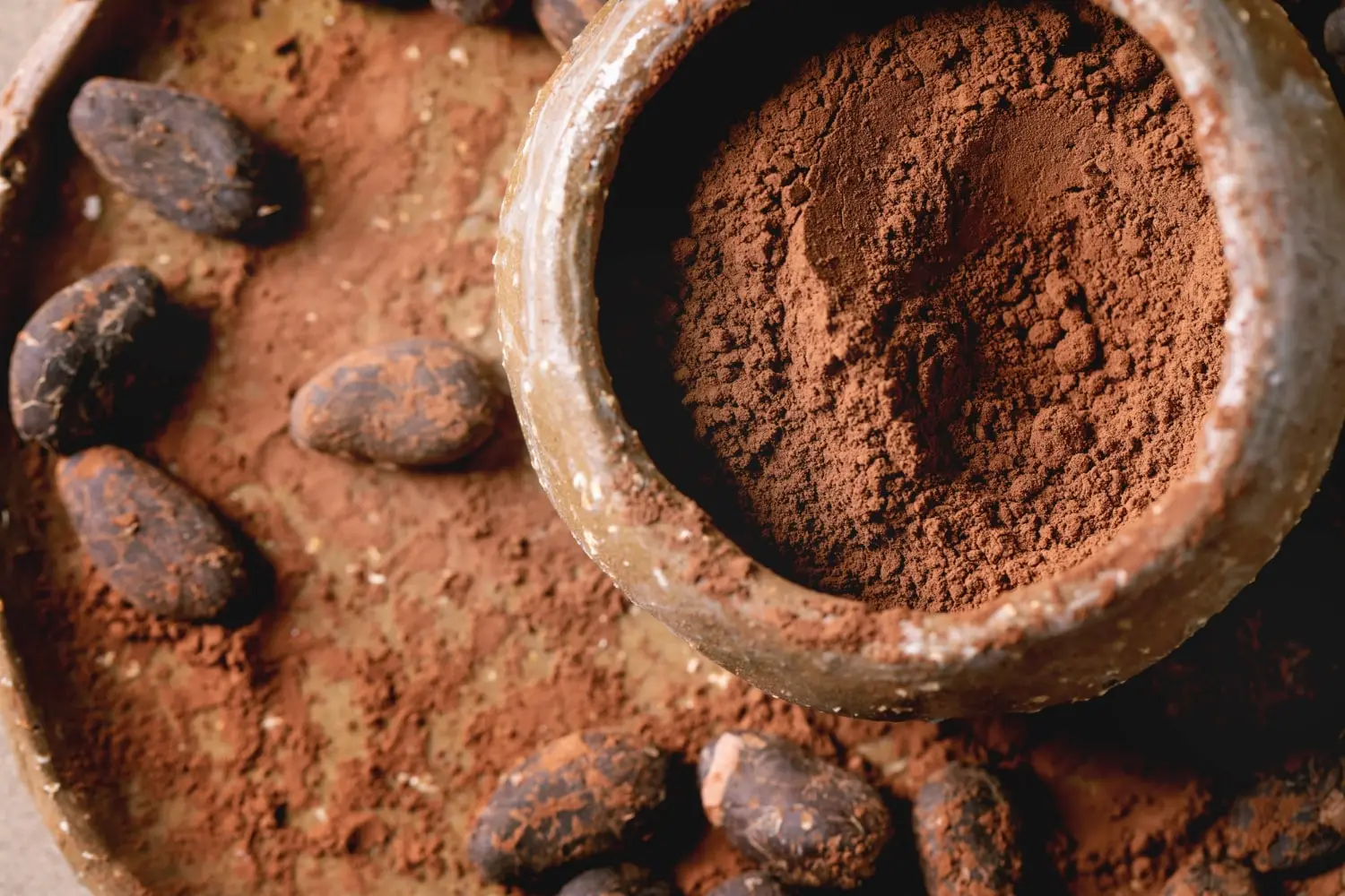 A imagem apresenta um close-up de um recipiente de cerâmica contendo cacau em pó. O recipiente está posicionado sobre uma superfície que parece ser de madeira ou pedra, e ao seu redor estão espalhados grãos de cacau inteiros.
