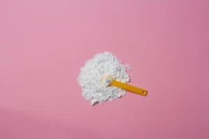 Imagem de uma porção de glutamina em pó branca, com uma colher medidora amarela sobre um fundo rosa claro.