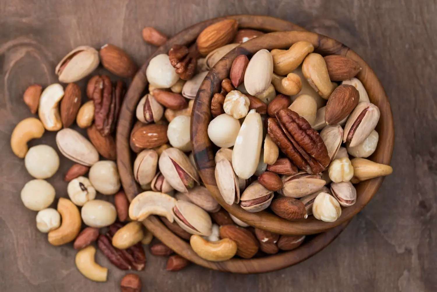 Imagem de uma variedade de oleaginosas em duas tigelas de madeira e espalhadas ao redor delas. As oleaginosas incluem castanhas, amêndoas, macadâmias, pecãs, pistaches e outras nozes, exibindo uma diversidade de cores e texturas. Imagem para o post "o que são oleaginosas"