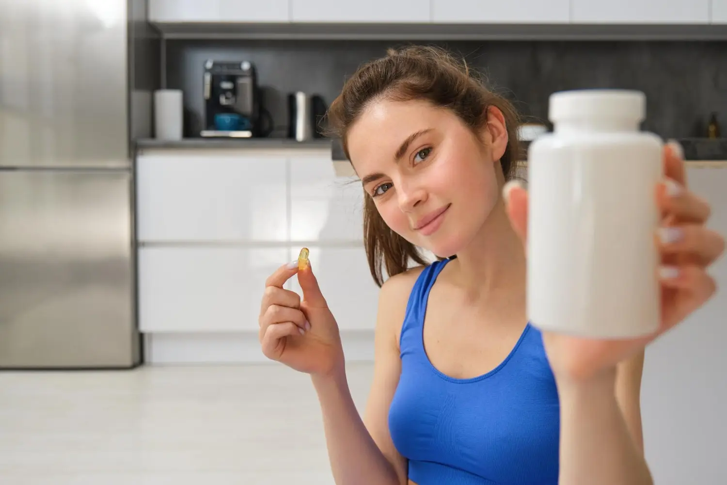 Mulher segurando uma cápsula de suplemento e um pote de BCAA, pronta para consumo. A cena retrata uma pessoa em ambiente doméstico, vestida com roupa de treino, destacando a praticidade do uso de suplementos para otimizar a recuperação muscular e melhorar o desempenho físico.