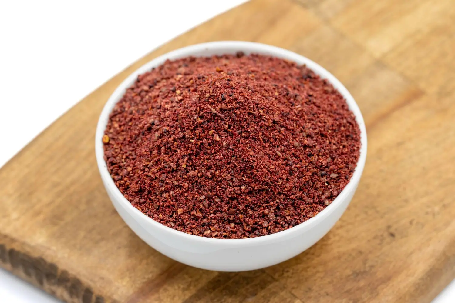 Tigela branca cheia de sumac em pó, um tempero vermelho escuro, posicionada sobre uma tábua de madeira clara.