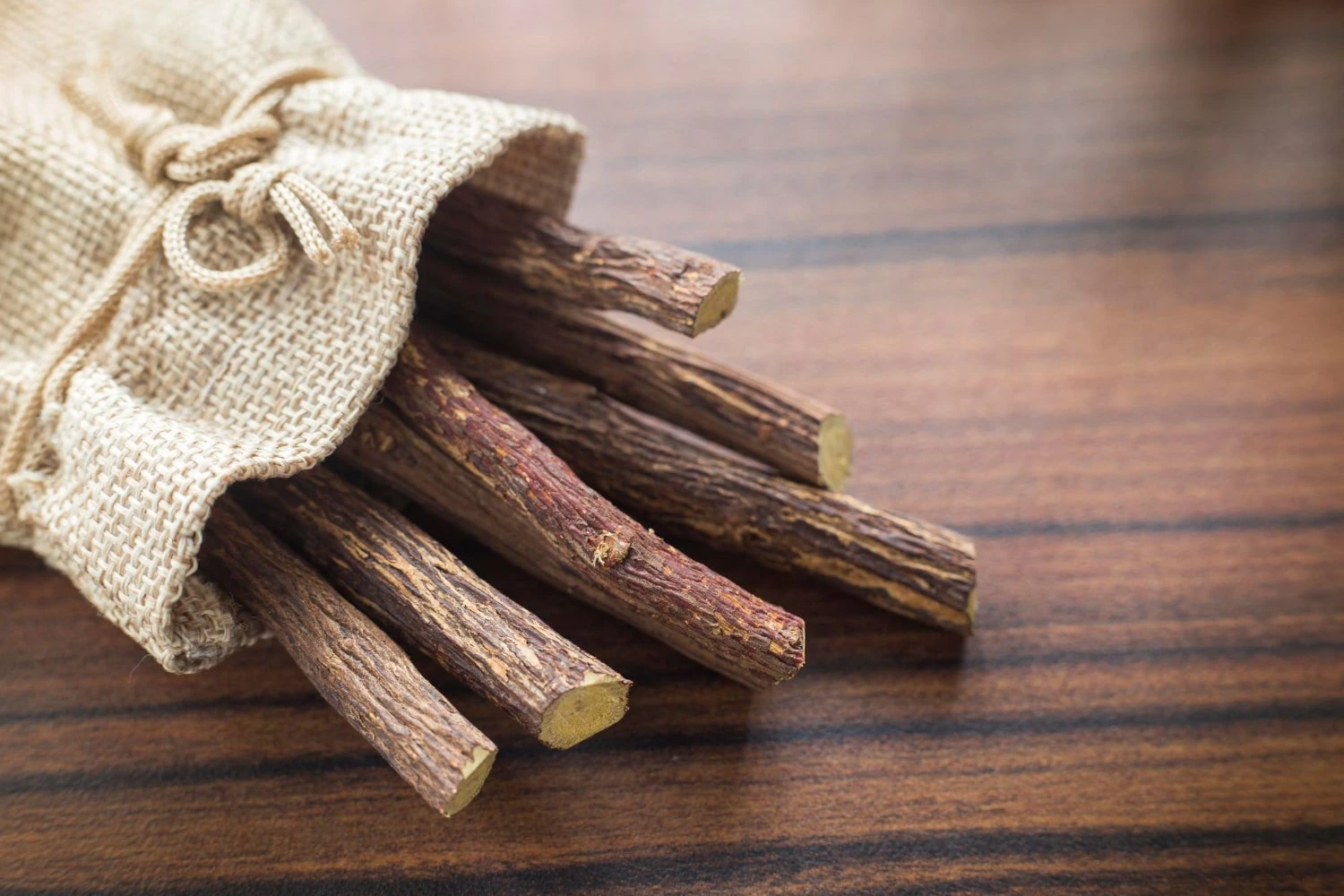 Várias raízes de alcaçuz dentro de um saco de pano bege, com detalhes rústicos em uma mesa de madeira. As raízes têm uma aparência marrom e texturizada.