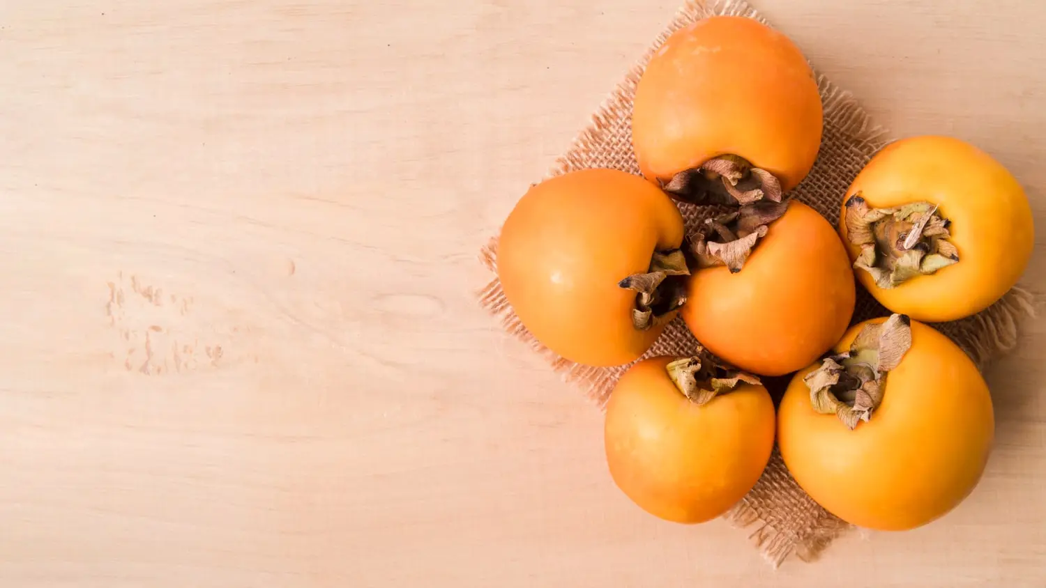 Seis caquis maduros dispostos sobre uma superfície de madeira clara, com uma base de tecido rústico sob as frutas. Os caquis apresentam uma cor laranja vibrante e folhas secas nas extremidades.