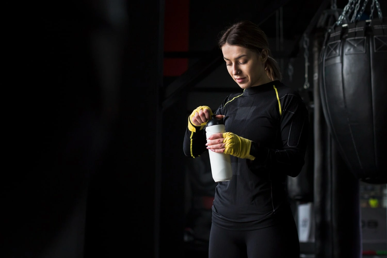 Mulher em uma academia, usando luvas de treino, segurando uma garrafa de suplemento enquanto se prepara para o treino, em um ambiente de fitness.