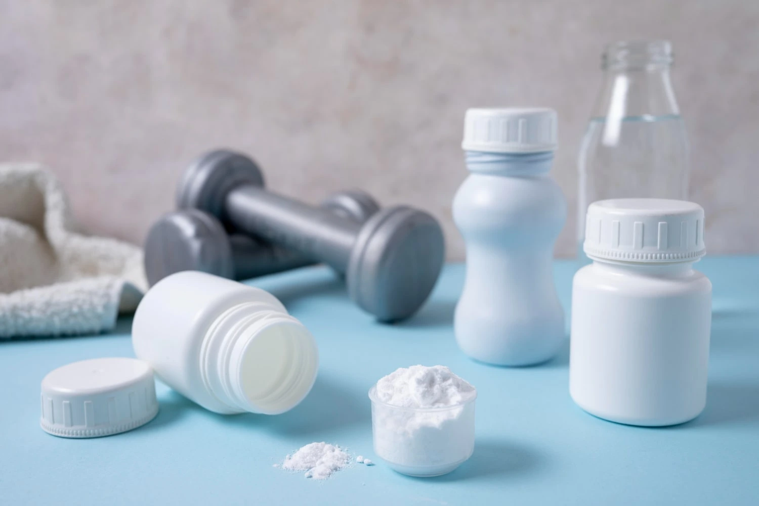 Frascos de suplementos com creatina em pó em destaque sobre uma superfície azul, ao lado de halteres e uma garrafa de água, simbolizando seu uso no ambiente de treino.