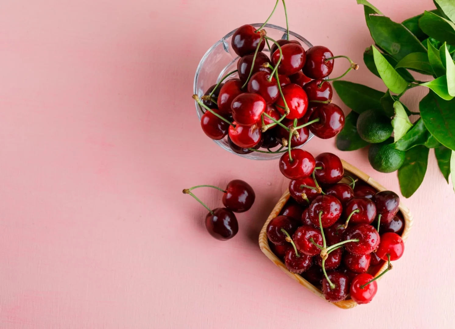 Cerejas frescas em uma tigela de vidro e uma cesta sobre uma superfície rosa, acompanhadas de folhas verdes ao lado.