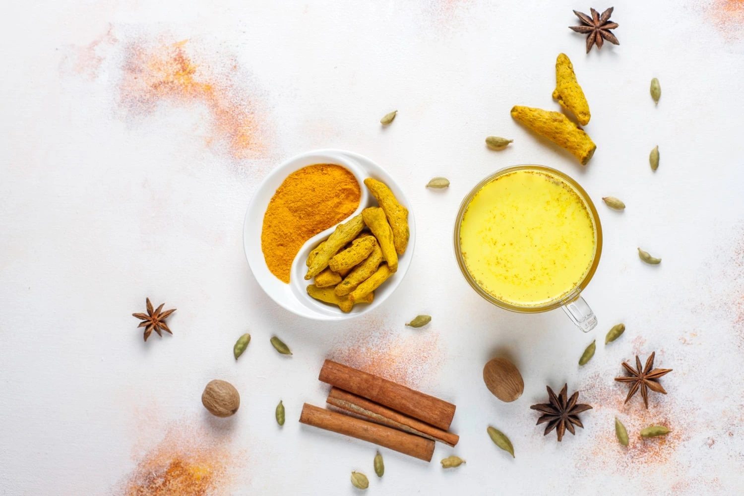 Golden milk servido em uma xícara transparente, cercado por ingredientes como cúrcuma em pó, raízes de cúrcuma, canela em pau, anis estrelado e cardamomo, sobre uma superfície clara.