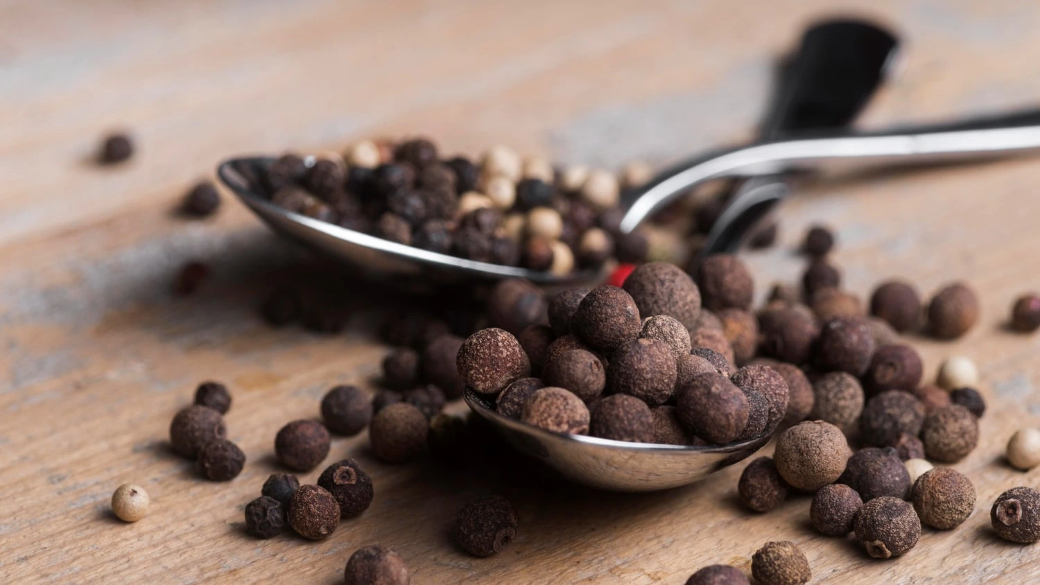Grãos de pimenta-da-jamaica espalhados em uma mesa de madeira, com duas colheres de metal contendo algumas das especiarias.
