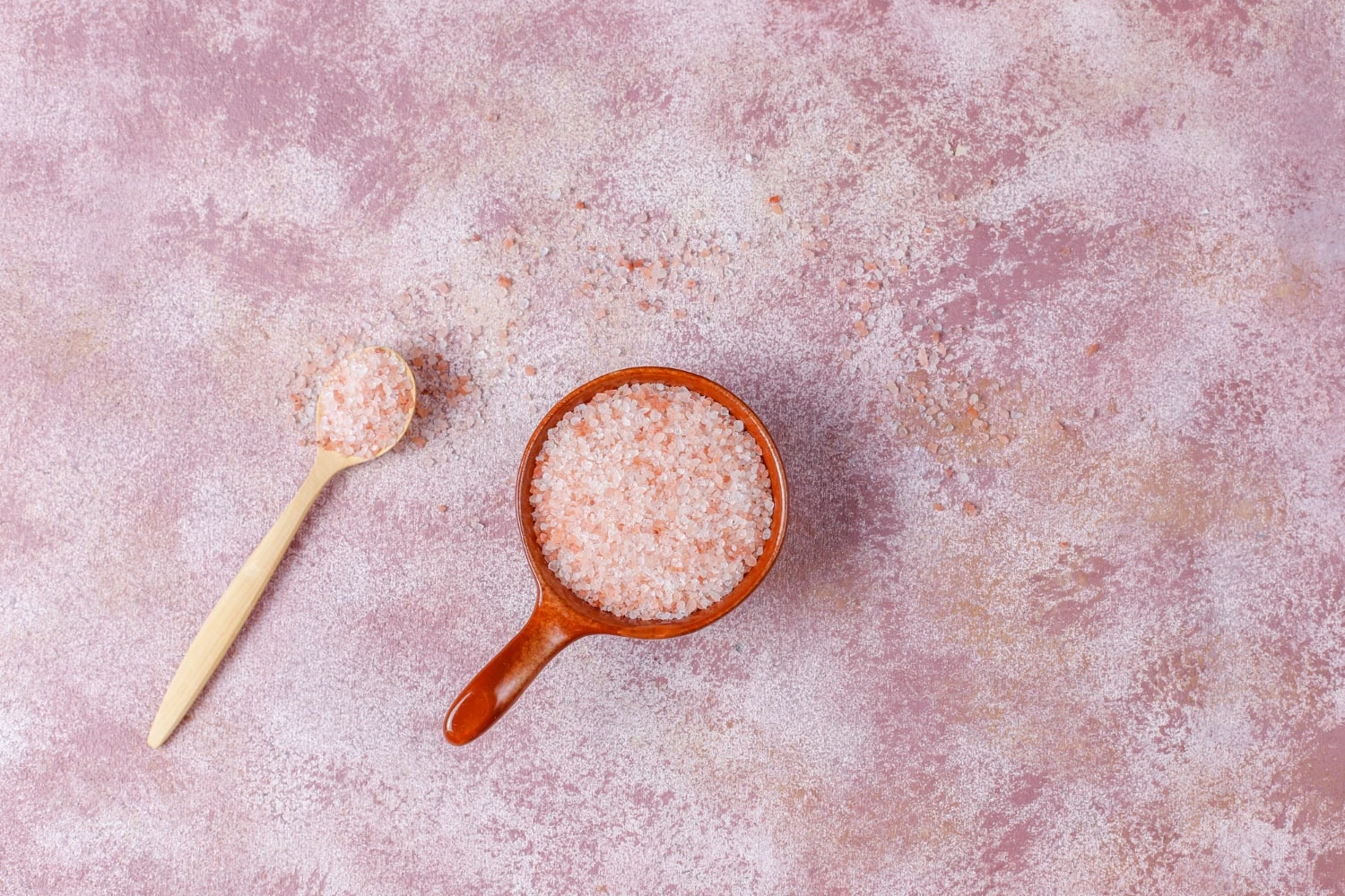 Sal rosa do Himalaia fino em uma colher de madeira ao lado de um recipiente de cerâmica, sobre uma superfície texturizada em tons de rosa.