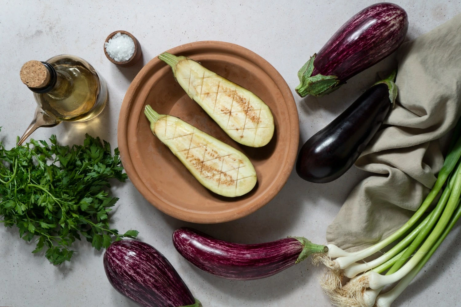 Berinjelas frescas em diferentes variedades dispostas ao lado de ingredientes naturais, como azeite, sal grosso, salsa e cebolinhas, com uma travessa de barro contendo berinjelas cortadas ao meio e marcadas para preparo, compondo uma cena de culinária saudável e rústica.