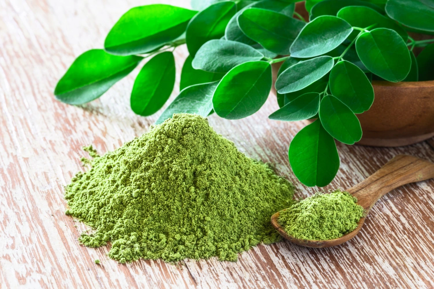 Folhas verdes frescas de moringa ao lado de uma porção de pó de moringa em tom verde vibrante, dispostos sobre uma superfície de madeira, destacando a textura natural do pó e das folhas.