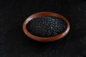 Foto de um recipiente de madeira com cristais de sal negro do Himalaia sobre uma superfície preta, destacando a textura e a cor escura do sal.